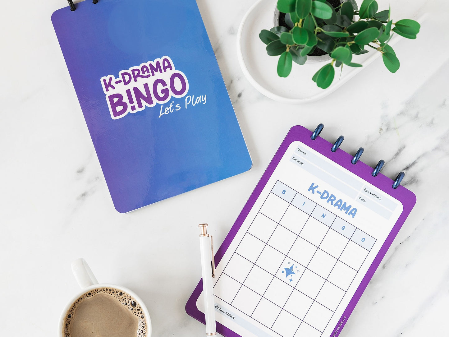 K-Drama Bingo Discbound Journal with one closed copy showing blue cover and one open copy showing blank purple bingo card on a marbled surface with a plant and coffee cup.