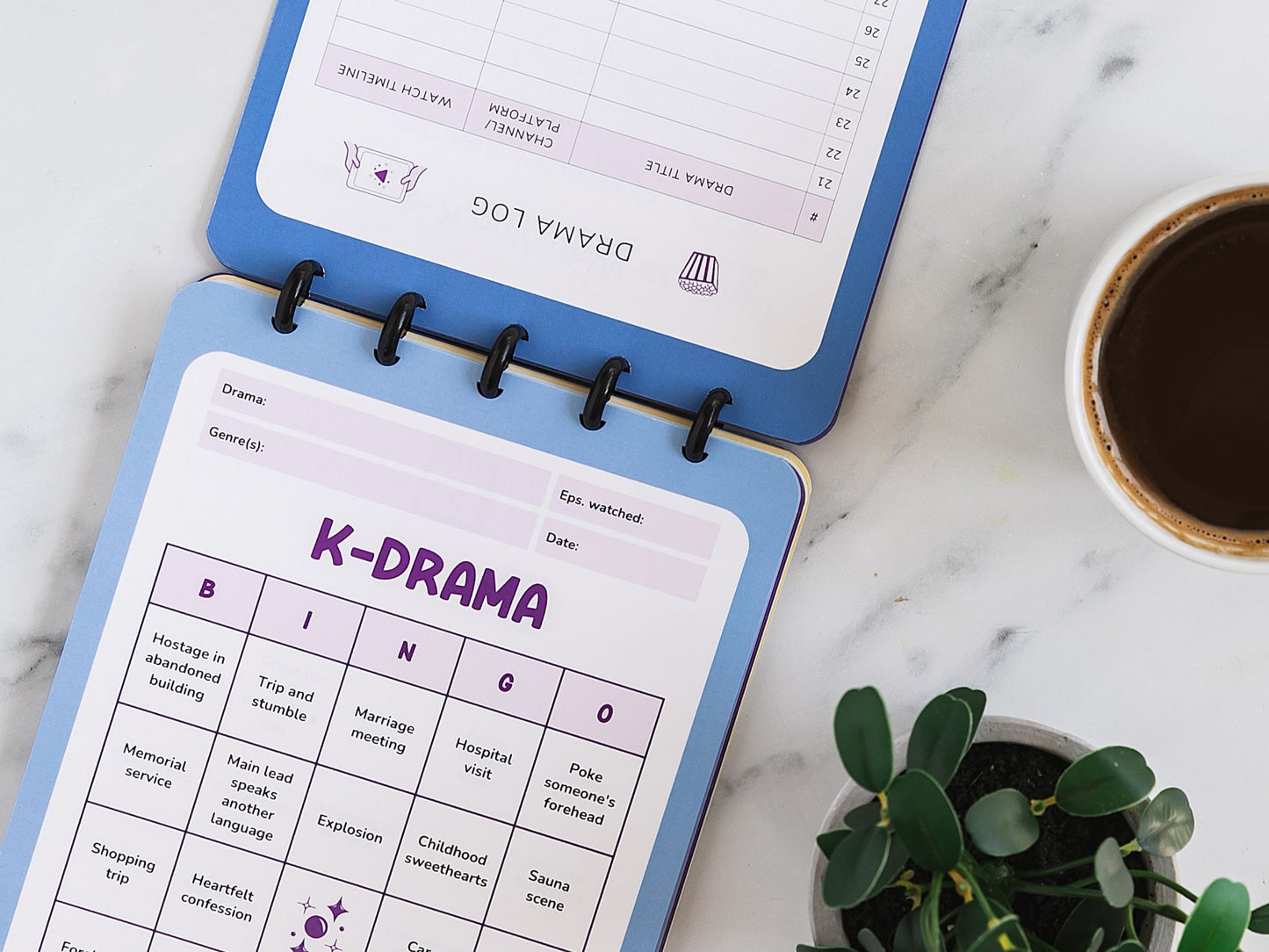 K-Drama Bingo Discbound Journal sitting open to a bingo card pre-filled with drama tropes on a marbled surface with a plant and coffee cup.