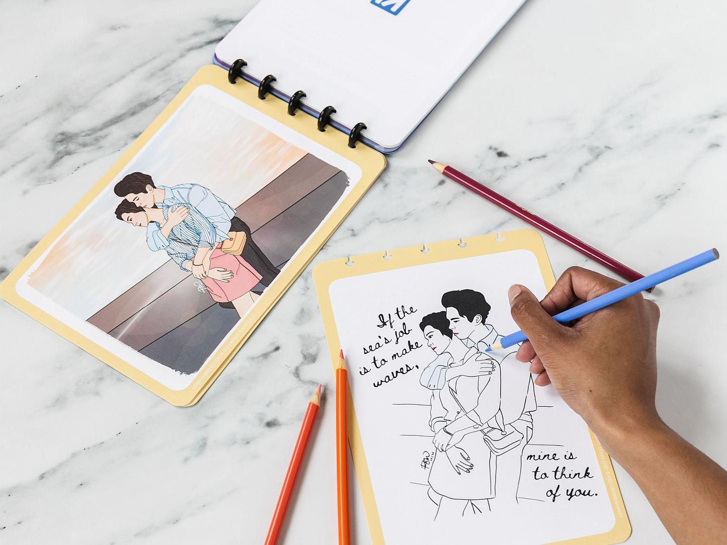 K-Drama Bingo Discbound open to show a person coloring a romantic illustration on coloring book page with colored pencils on a marble surface.